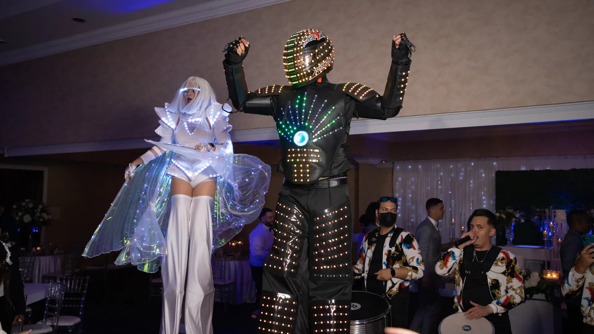 Robot LED y zanquera con alas de luz junto a músicos en un salón