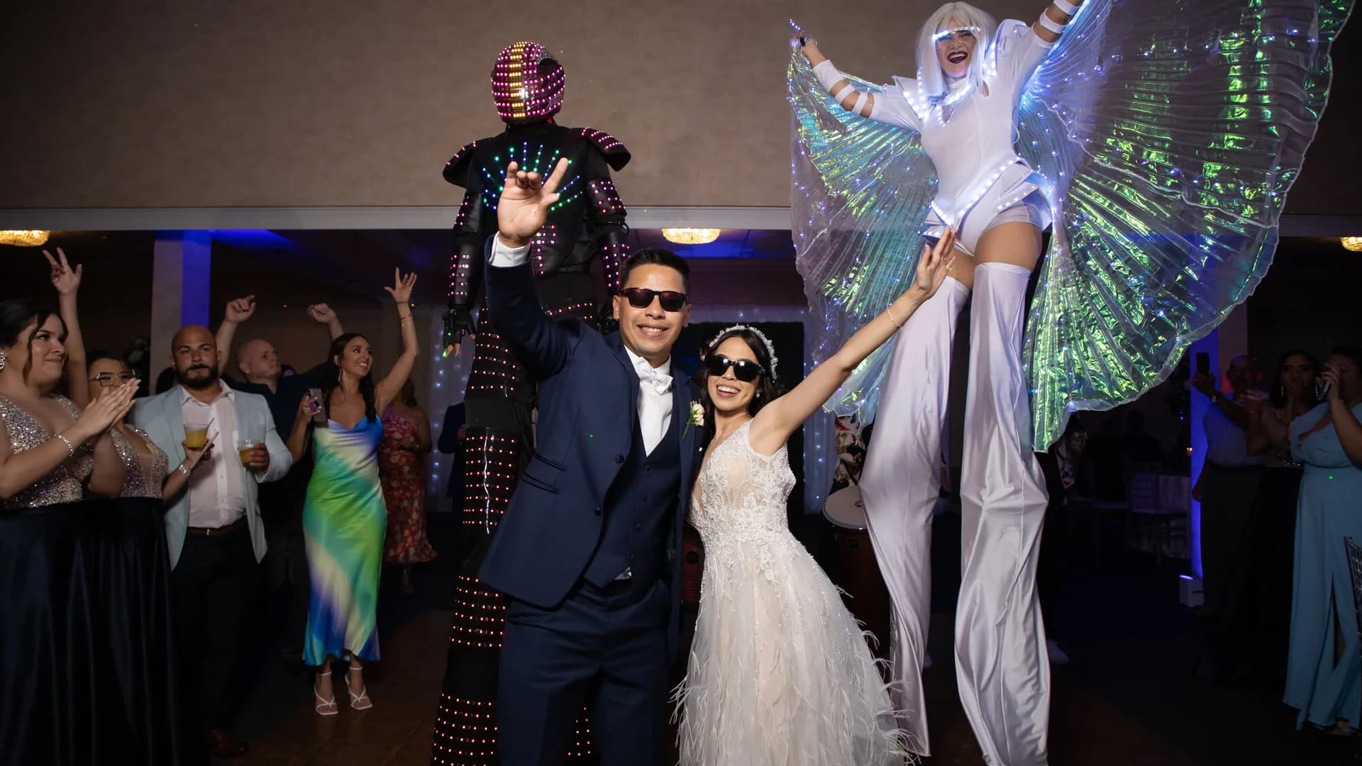 Novios con gafas oscuras junto a un robot LED y una zanquera con alas de luz