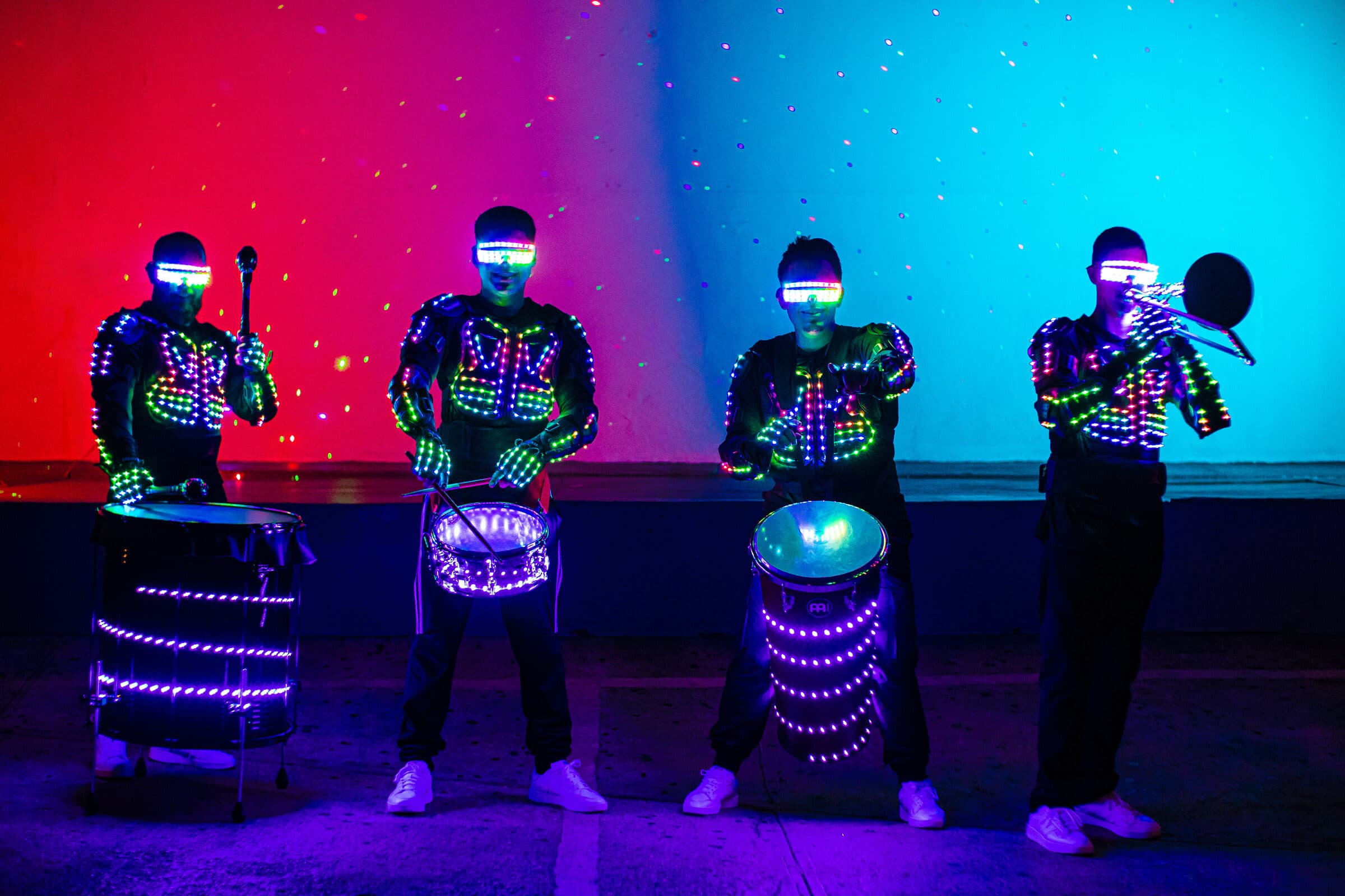 Batucada LED con trajes y tambores iluminados en azul y morado sobre fondo rojo y turquesa