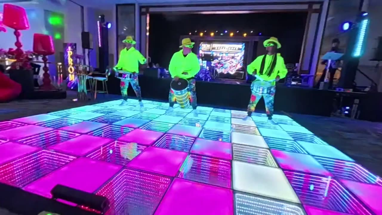 Tres drummers con vestuario neón tocando sobre una pista iluminada