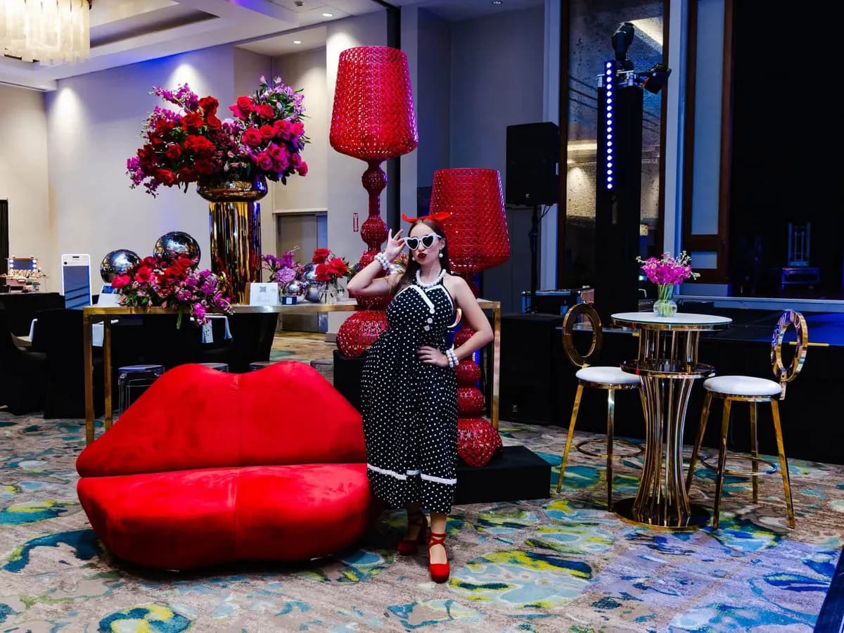 Personaje pop con vestido de lunares y gafas blancas junto a un sofá en forma de labios