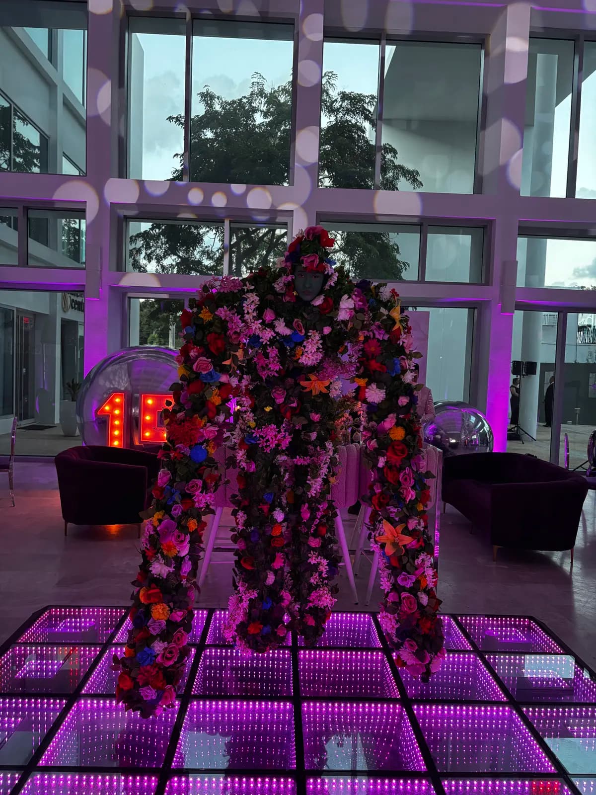 Personaje cubierto de flores de colores de pie frente a una pista LED 3D infinity rosa en un salón con ventanales