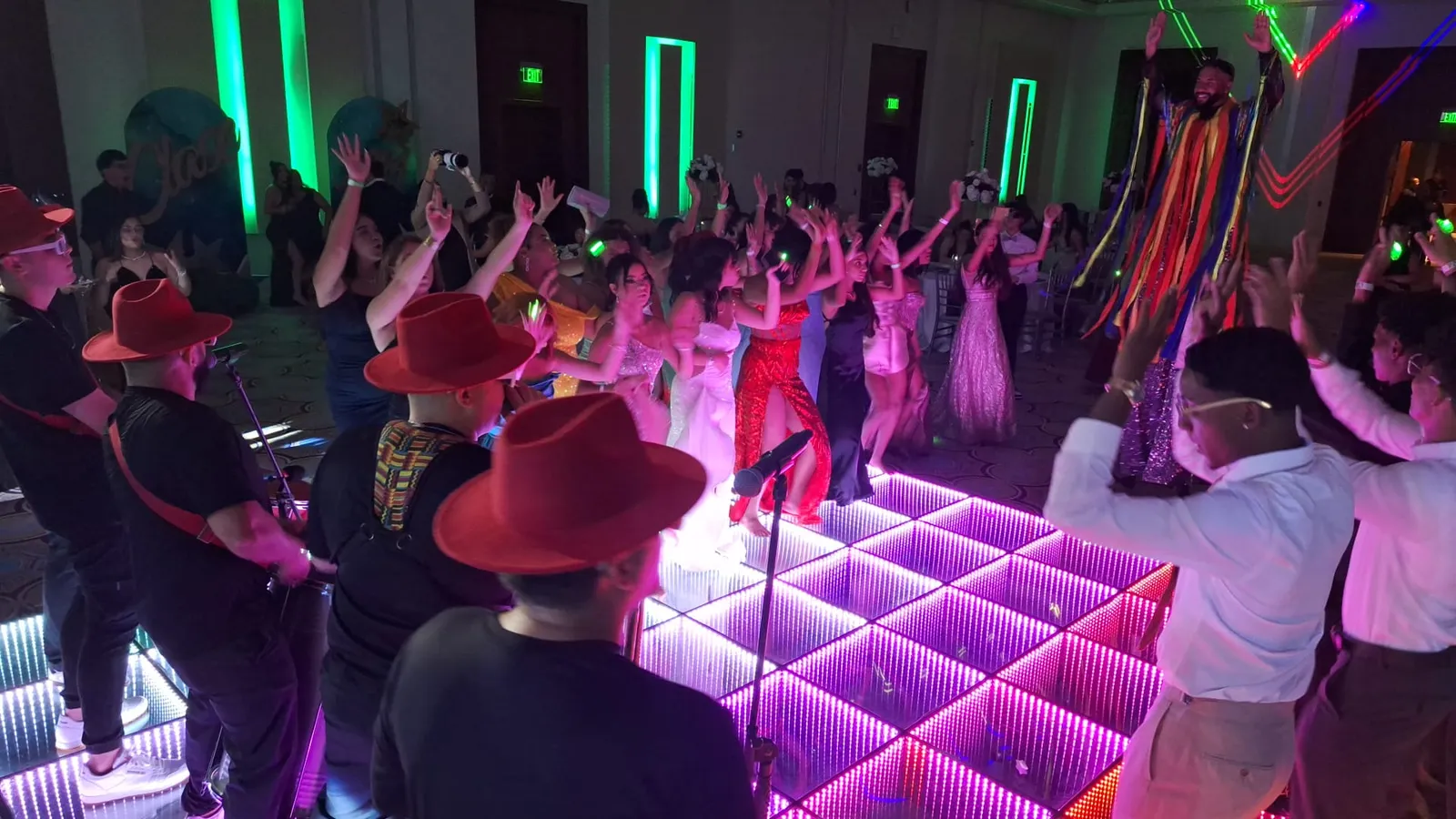 Invitados con los brazos en alto bailando sobre una pista LED rosa junto a una batucada de sombreros rojos