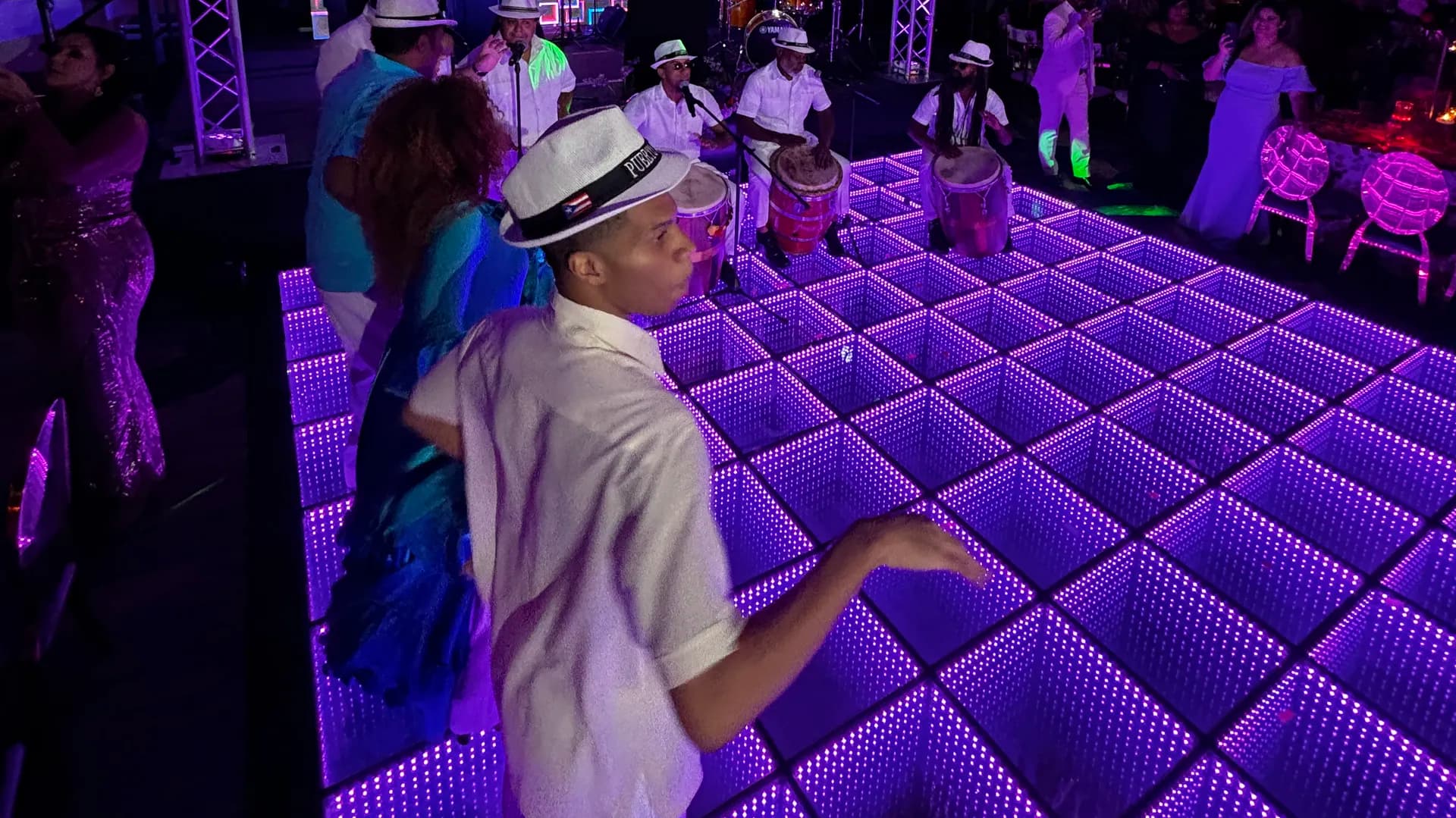 Pista de baile LED 3D infinity morada con efecto espejo y músicos de batucada de camisa blanca y sombrero tocando al fondo