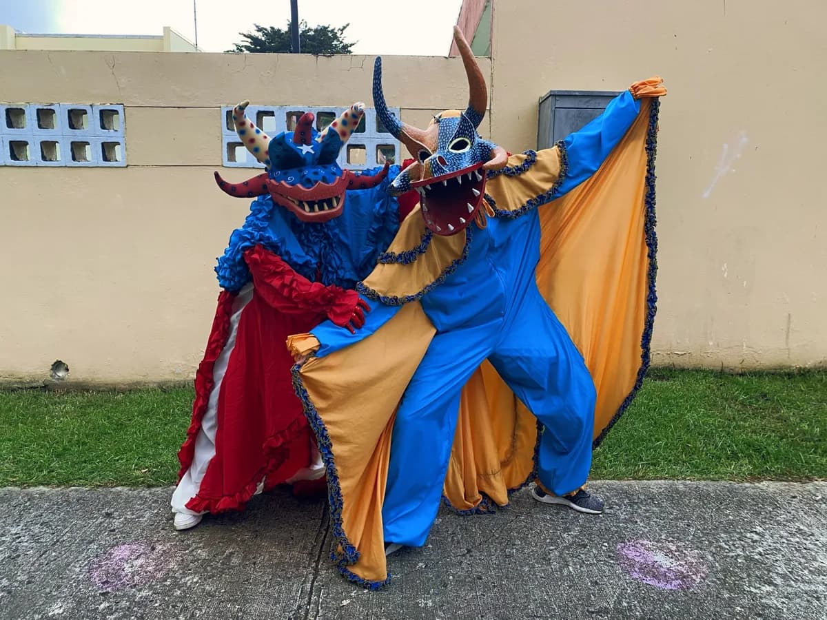 Dos vejigantes de piso con máscaras de cuernos y capas en azul, rojo y amarillo posando al aire libre