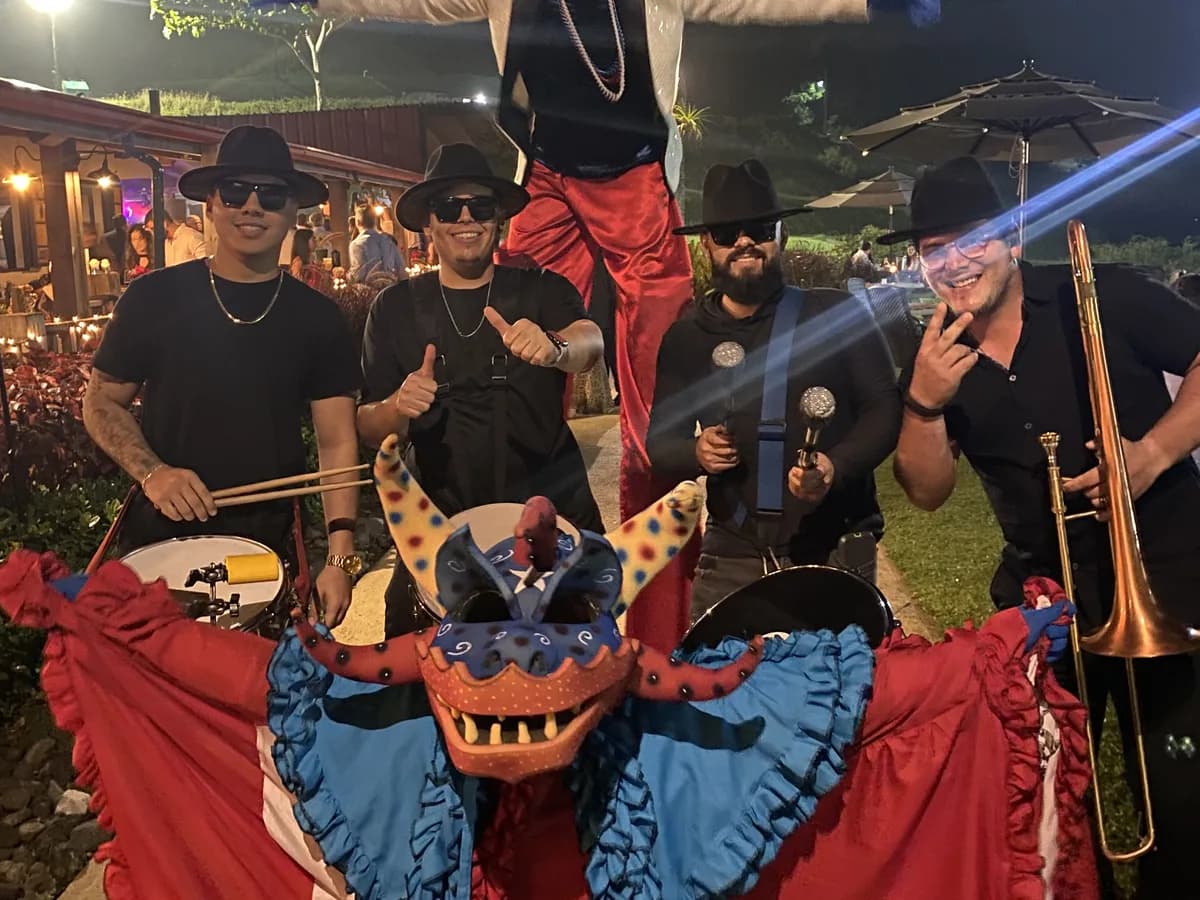 Vejigante con traje de la bandera frente a una batucada con sombreros y un zanquero de brazos abiertos