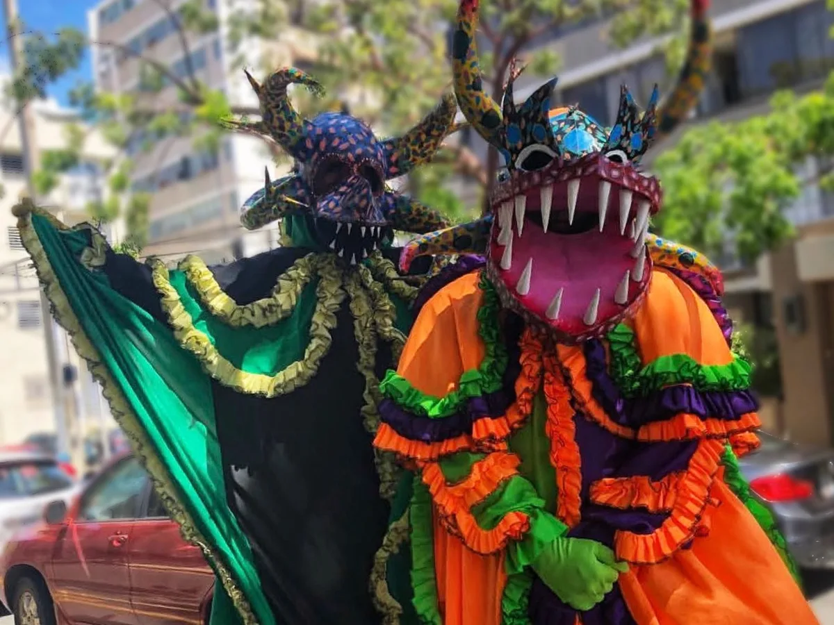 Dos vejigantes con trajes de volantes en verde, morado y naranja posando en una calle soleada