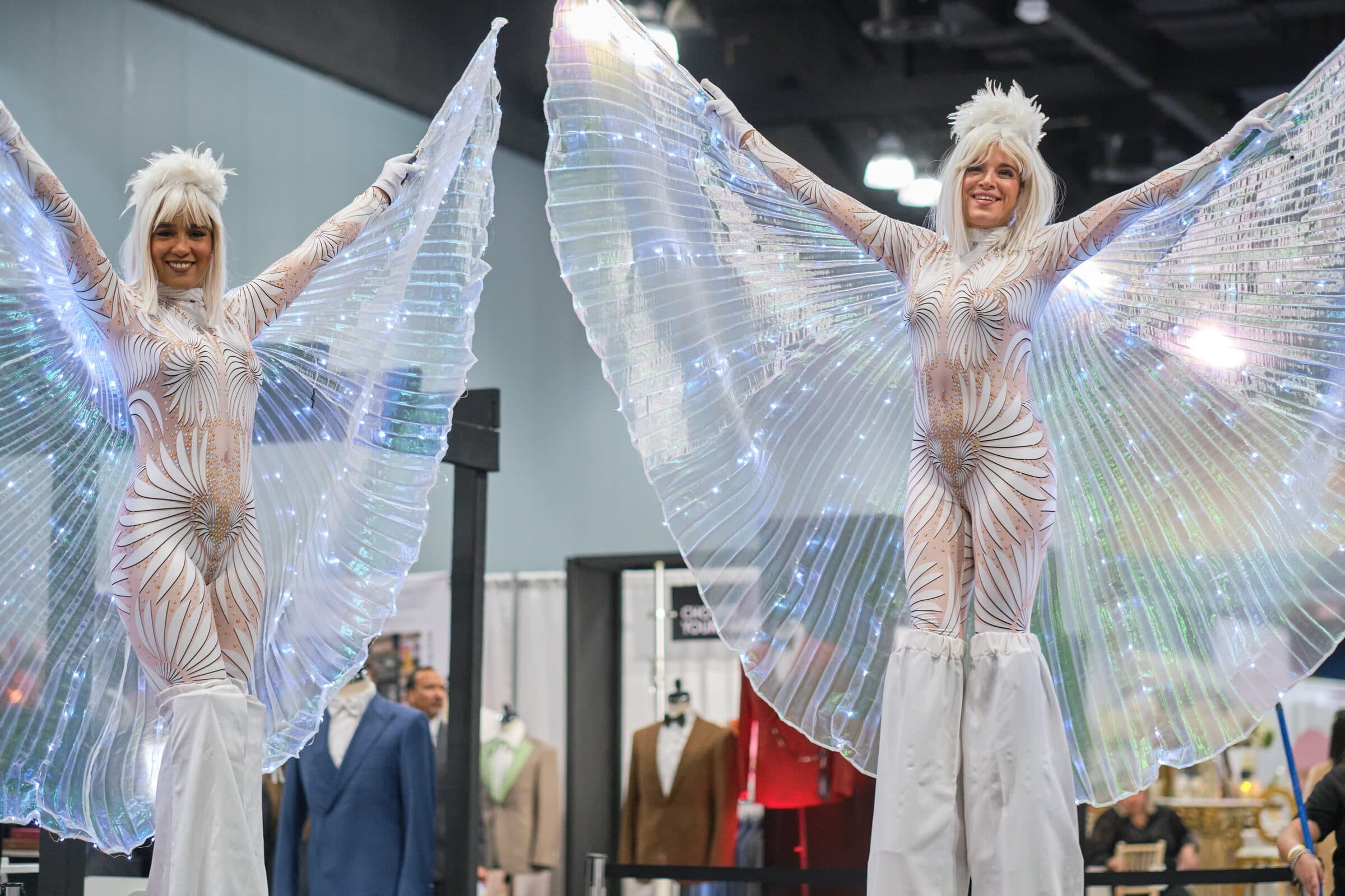 Dos zanqueras con alas doradas iluminadas recorriendo una feria de bodas
